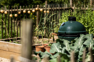 A green ceramic kamado grill sits in a sunny garden plot with leafy greens, potted plants, and a wooden fence in the background.