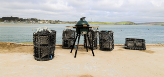 A black ceramic kamado grill on a tripod stands on a sandy shore with ocean water and distant hills in the background. Several black mesh lobster traps are arranged around it.