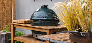 A black ceramic kamado grill sits on a wooden table in an outdoor patio setting, with pampas grass in a pot in the foreground and a gray wall behind.